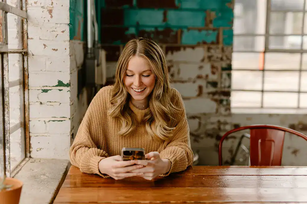 Women sitting at a table wearing a brown sweater smiling at her phone